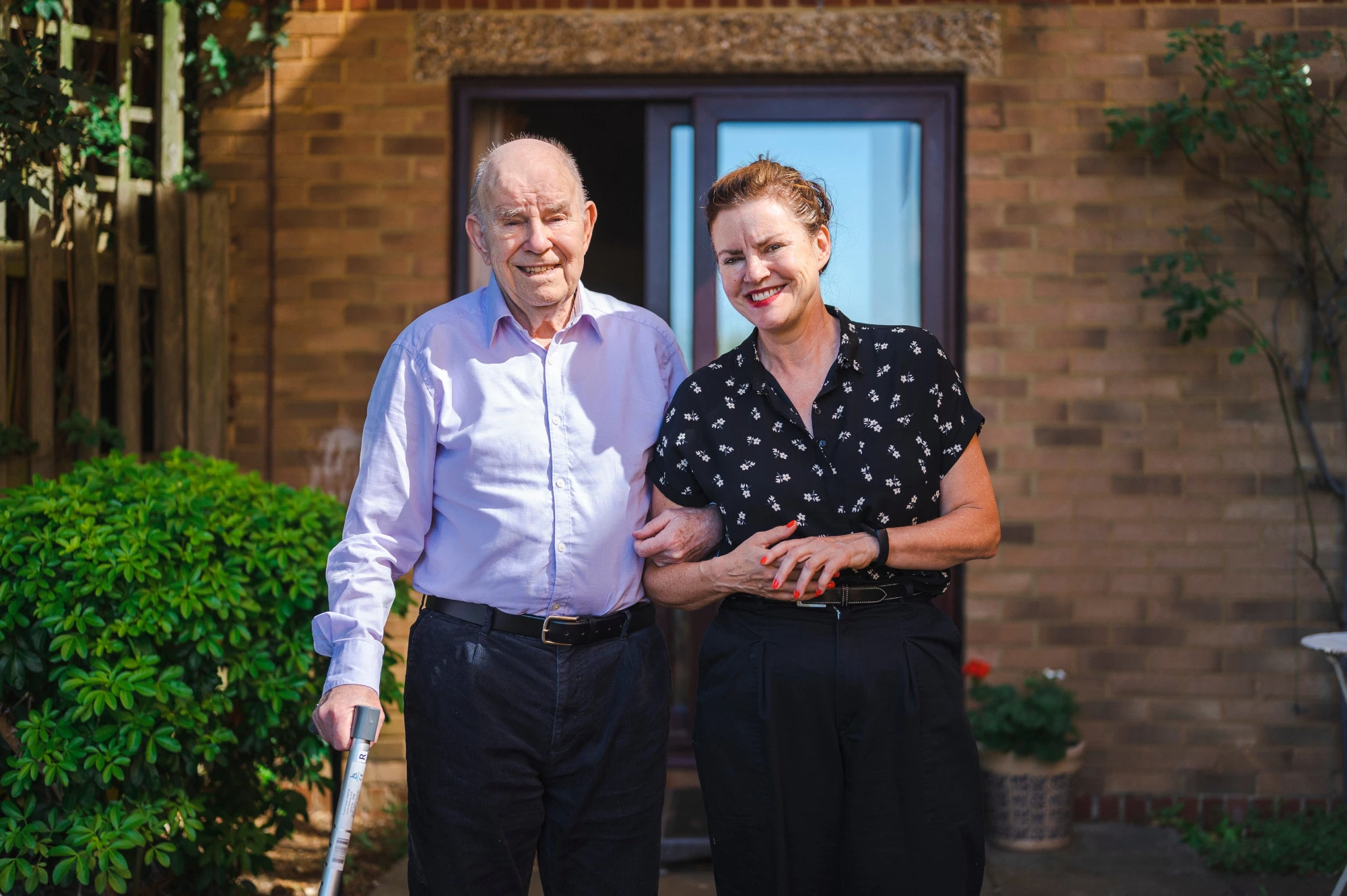 Woman linking arms with older man to support his walking in the garden