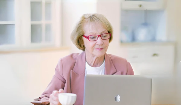 Older Woman With A Cup Of Tea Looking At Laptop On Kitchen Table